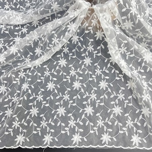 Op de afbeelding: Witte bloemen geborduurde stof. Het transparante materiaal heeft een herhalend patroon van delicate witte bloemen en wijnranken. De stof is lichtgewicht en heeft een geschulpte rand, geschikt voor diverse knutsel- en designprojecten.