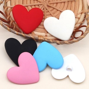 Puede incluir: Una colección de adornos en forma de corazón en varios colores, incluyendo rojo, blanco, negro, rosa y azul claro. Los corazones están dispuestos sobre y alrededor de una cesta tejida. Estos corazones decorativos son adecuados para manualidades y proyectos de bricolaje.