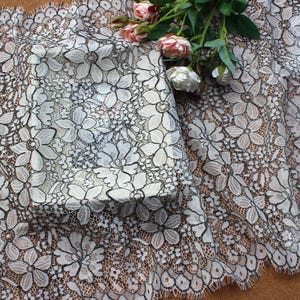 Peut inclure: Tissu en dentelle florale ivoire avec une bordure festonnée. La dentelle présente un motif répété de grandes fleurs stylisées avec des contours noirs. Quelques roses roses et blanches sont posées sur le tissu.