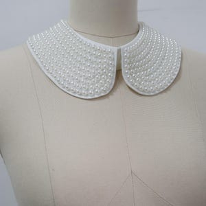 Może przedstawiać: Biały, odpinany kołnierz gęsto pokryty małymi, okrągłymi, sztucznymi perłami. Kołnierz jest w klasycznym stylu Piotrusia Pana, z zaokrąglonymi krawędziami i lekkim zakrzywieniem, aby dopasować się do dekoltu. Perły są jednolite.