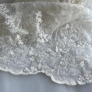 Può includere: Tessuto di pizzo color avorio con motivo floreale. Il delicato pizzo presenta fiori e foglie ricamate, con un bordo smerlato. Il tessuto è semitrasparente, lasciando passare la luce, e viene probabilmente utilizzato per abiti da sposa o altri capi per occasioni speciali.