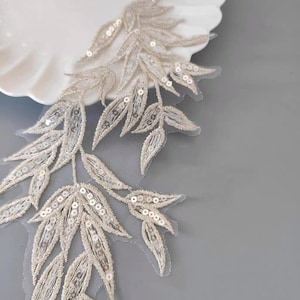 May include: A delicate, leaf-shaped applique with intricate detailing. The leaves are embellished with shimmering sequins and a subtle, neutral color palette. The applique is likely for embellishing garments or craft projects.