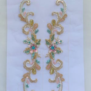 May include: A pair of symmetrical floral appliques featuring intricate beadwork and sequins in shades of gold, soft pink, mint green, and bright blue. The elongated, curved designs are embellished with white seed beads and metallic gold thread.