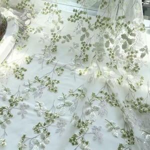 May include: A sheer white tulle fabric featuring an intricate embroidered pattern of delicate branches with leaves and small clusters of berries. The design is embellished with shimmering light green and silver sequins that catch the light.