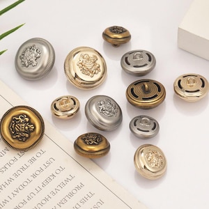 May include: A collection of round metal buttons in gold and silver tones, featuring embossed heraldic crest designs and textured edges. Some buttons have a smooth finish, while others display a perforated pattern. Text is visible on the paper.