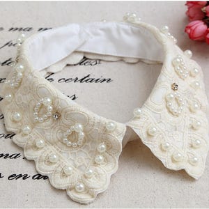 Puede incluir: Cuello de encaje marfil adornado con perlas de imitación y pequeños pedrería. El cuello presenta un borde festoneado y un reverso de tela blanca. Este elegante accesorio añade un toque de encanto vintage a cualquier atuendo.