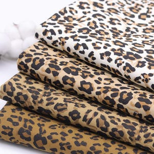 May include: Close-up of stacked fabric with a leopard print pattern. The fabric features a tan background with dark brown and black spots. The fabric is folded, showing multiple layers of the animal print.
