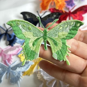 May include: A green embroidered butterfly patch with different shades of green. The butterfly has detailed wing patterns and is held in a hand. Other colourful butterfly patches are visible in the background, including blue, pink, and orange.
