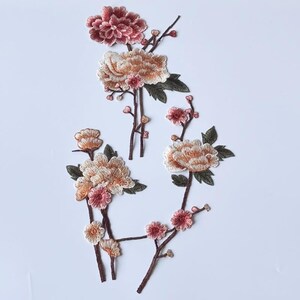 May include: Three embroidered floral patches featuring blooming peonies and smaller buds in shades of dusty rose, cream, and peach. The designs include dark brown stems and deep green leaves, rendered with detailed thread work on a plain background.