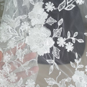 Puede incluir: Primer plano de tela de encaje floral blanco. El delicado encaje presenta flores bordadas, hojas y peque&ntilde;as lentejuelas. La tela es transparente, con un fino fondo de malla. Ideal para ropa de novia o de ocasi&oacute;n especial.