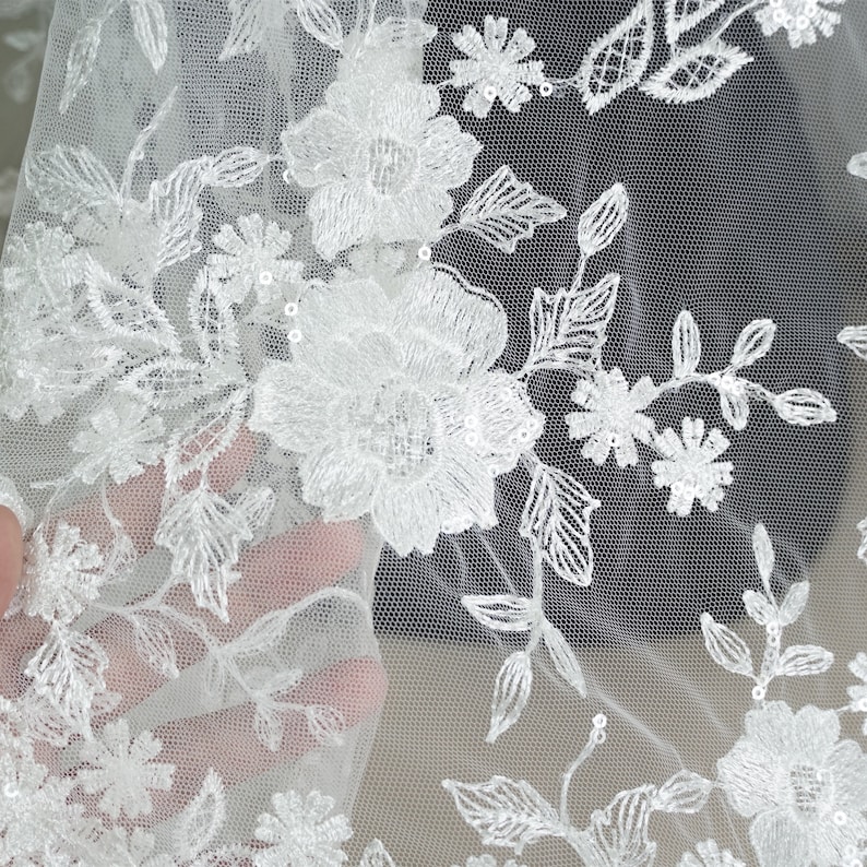 Puede incluir: Primer plano de tela de encaje floral blanco. El delicado encaje presenta flores bordadas, hojas y peque&ntilde;as lentejuelas. La tela es transparente, con un fino fondo de malla. Ideal para ropa de novia o de ocasi&oacute;n especial.