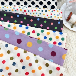 May include: A stack of folded fabric pieces featuring various polka dot patterns. The fabrics include white with black dots, white with multicoloured dots, black with neon dots, cream with colourful dots, lavender with large dots, and white with red and gold dots.
