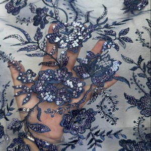 Puede incluir: Tela de encaje floral azul marino con intrincados detalles de lentejuelas. El diseño presenta flores y hojas bordadas, creando un patrón delicado y texturizado. La tela es semitransparente.