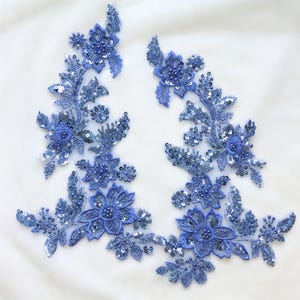 May include: Two blue floral appliques with detailed bead and sequin embellishments. The appliques feature various sizes of flowers and leaves, creating an ornate design. The embellishments are arranged on a sheer, white background.