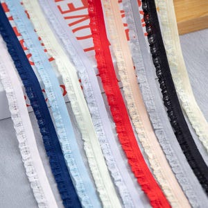 May include: A collection of colorful lace ribbons in various shades, including white, navy blue, light blue, red, and black. Each ribbon features a delicate ruffled edge, creating a textured and decorative element. The ribbons are arranged closely together, showcasing their diverse colors and textures.