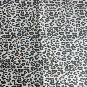 May include: A fabric with a leopard print pattern. The pattern features black and brown spots of varying sizes on a cream-colored background. The fabric has a smooth texture and a repeating design.