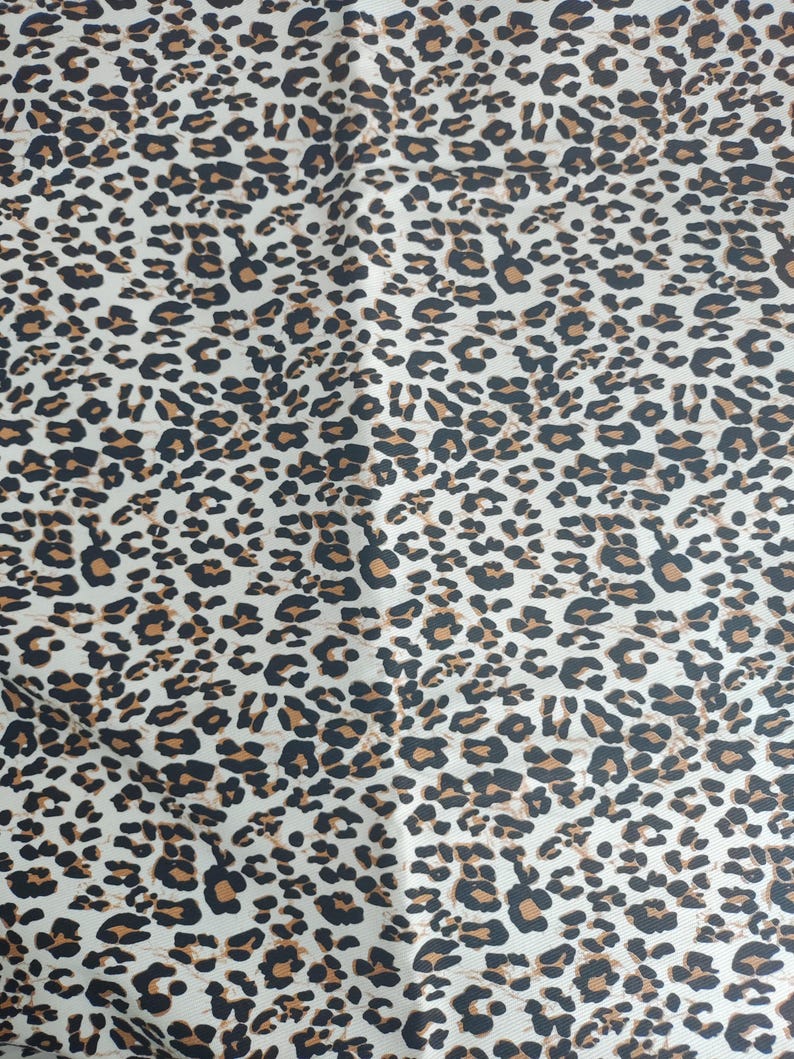 May include: A fabric with a leopard print pattern. The pattern features black and brown spots of varying sizes on a cream-colored background. The fabric has a smooth texture and a repeating design.