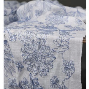 May include: White fabric with a blue floral embroidery design. The embroidery features large, detailed flowers and leaves. The fabric appears to be linen or a similar lightweight material, suitable for home decor or textiles.
