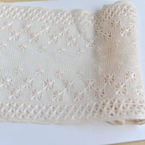 以下が含まれることがあります： クリーム色のレース生地が巻かれたもので、花柄と幾何学模様が繰り返されています。 レースは繊細でオープンな織り方で、詳細な刺繍が施されており、質感のある表面を作り出しています。 この生地は、手芸や装飾目的で使用される可能性があります。