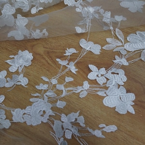 May include: White floral lace fabric featuring a delicate pattern of flowers and leaves. The lace is sheer and has a soft, flowing texture.