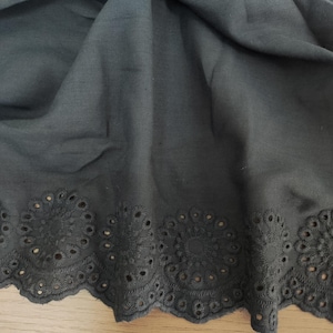 May include: Dark gray fabric with a scalloped eyelet lace trim. The fabric is draped, showing the texture and detail of the eyelet design. The eyelet pattern features circular floral motifs.