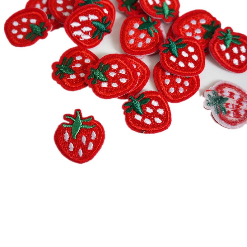 May include: A collection of red strawberry patches with green stems and white seeds. These embroidered patches are scattered across a white background. The patches are a vibrant red color.