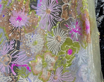 May include: Close-up of a sheer fabric with floral embroidery. The design features large flowers in shades of pink, gold, and green, embellished with sequins. The background is a light, neutral color, highlighting the colorful embroidery.