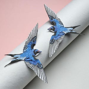 May include: Two embroidered swallow patches featuring vibrant blue bodies, black heads with red accents, and wings detailed with white and black stitching. The birds are depicted in flight with wings spread wide, showcasing intricate thread work and textures.