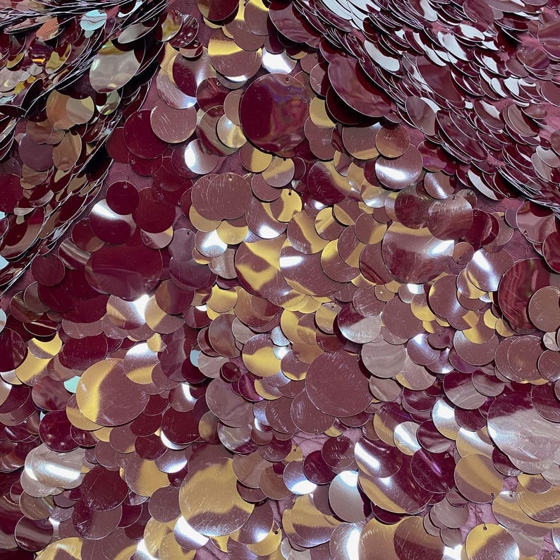 Puede incluir: Una superficie de tela densa cubierta de lentejuelas circulares superpuestas en un color burdeos intenso. Las lentejuelas var&iacute;an en tama&ntilde;o y presentan un acabado brillante y reflectante que crea luces y sombras en el material.