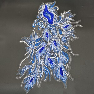 Può includere: Un'applicazione decorativa con un motivo a piuma di pavone ricamato con filo blu vibrante e argento. Il pezzo è impreziosito da paillettes blu scintillanti e accenti metallici argentati, disposti in un motivo fluido su una base in rete trasparente.