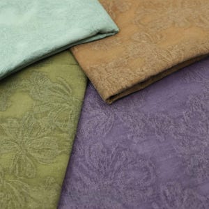 May include: Close-up of four fabric swatches in shades of green, brown, and purple. Each swatch has a floral pattern embossed on the surface. The fabrics are folded, revealing their textures and colours.