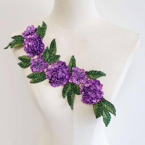 May include: A decorative floral applique featuring vibrant purple sequined flowers and green beaded leaves. The applique is designed in a cascading arrangement, perfect for embellishing garments or accessories. The flowers have a layered petal design.