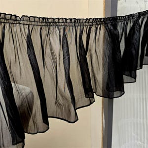 Puede incluir: Un trozo de tela negra transparente con un borde con volantes. La tela está fruncida en la parte superior, creando un efecto fluido y en capas. El material es translúcido, permitiendo el paso de la luz, y probablemente se utiliza para ropa o fines decorativos.