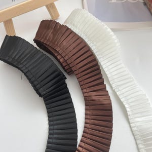 Puede incluir: Primer plano de tres cintas plisadas en negro, marrón y blanco. Las cintas están dispuestas una al lado de la otra, mostrando su diseño texturizado y plegado. La tela parece ser satén o un material similar, con un ligero brillo.