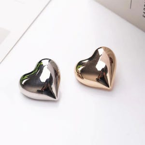 May include: Two heart-shaped decorative objects. One is silver, the other is gold. Both have a smooth, reflective surface. The hearts are placed on a white surface, suggesting a minimalist aesthetic. The objects are likely used for decoration or as a gift.