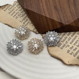 Może przedstawiać: Cztery kolczyki sztyfty w kształcie kwiatów. Każdy kolczyk ma centralny przezroczysty kamień otoczony kamieniami o szlifie bagietowym. Dwa są w kolorze srebrnym, a dwa w kolorze złotym, prezentowane na białej powierzchni.