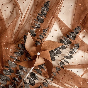 May include: A brown paper pinwheel with a white stick, surrounded by dried eucalyptus branches on a brown tulle fabric with gold glitter stars.