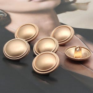 Puede incluir: Colección de seis botones dorados. Los botones son redondos con un centro en relieve y un borde decorativo y acanalado. Un botón está volteado, revelando el lazo en la parte posterior.