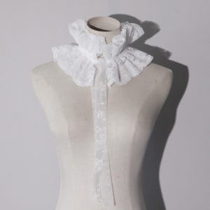 May include: A white lace detachable collar featuring a high, ruffled neckline with a delicate floral pattern. The collar includes two long, thin lace ribbons that hang vertically from the center front, creating a layered and textured appearance.
