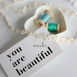 May include: White beaded trim with a scalloped edge. The trim is on a white surface with a card that says "you are beautiful".