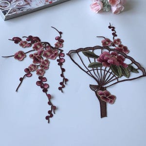 May include: Embroidered floral appliques in shades of burgundy, pink, and green. One applique is a branch of flowers, the other is a fan shape with flowers and leaves. These decorative appliques can be used for various craft projects.