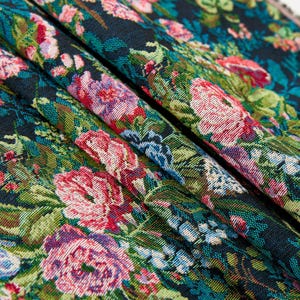 Puede incluir: Una tela tejida con un denso patr&oacute;n floral de flores de color rosa vibrante, rojo y azul claro sobre un fondo negro oscuro. El tejido muestra un intrincado follaje y hojas verdes, creando un dise&ntilde;o bot&aacute;nico texturizado y multicolor.