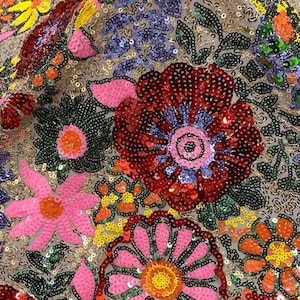 May include: Close-up of a fabric with a floral design. The fabric is covered in sequins in various colors, including red, pink, yellow, and blue. The flowers are arranged in a repeating pattern on a beige background.
