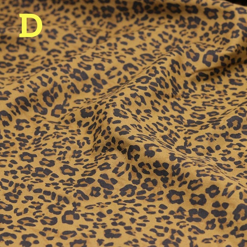 May include: Mustard-colored fabric with a leopard print pattern. The fabric has a repeating design of dark brown spots on a tan background. The letter "D" is in the upper left corner of the image.