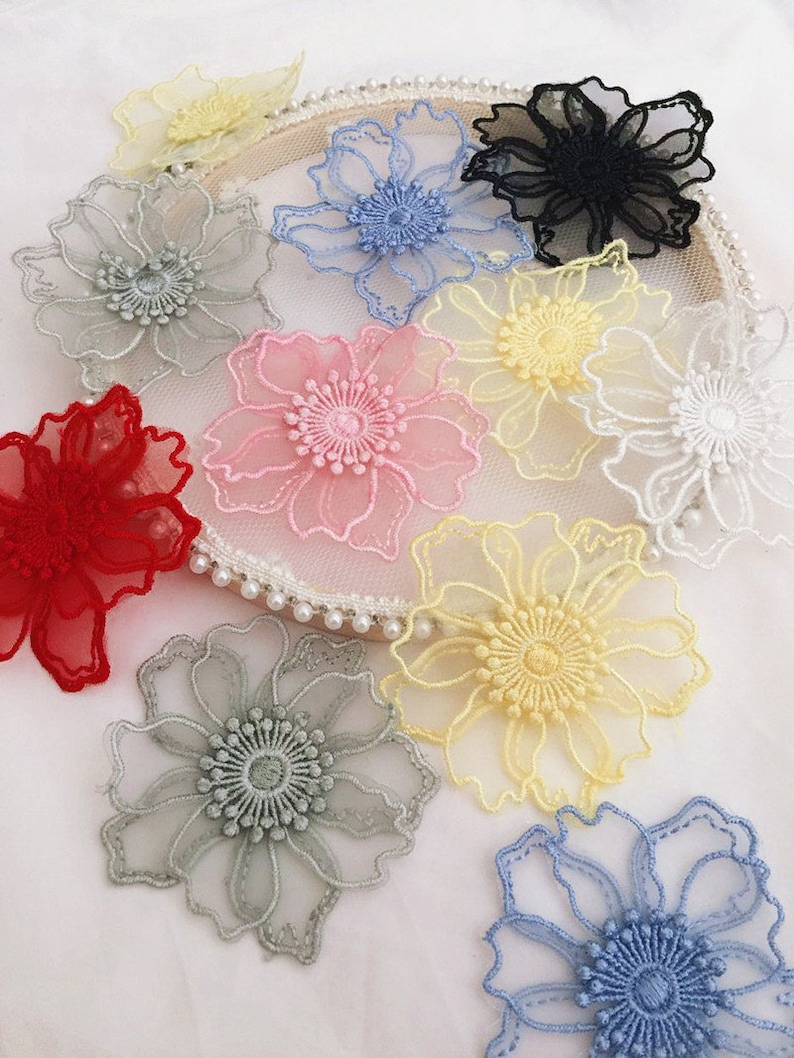 May include: A collection of multi-colored floral lace appliques in shades of red, pink, yellow, white, blue, gray, and black. Each flower features intricate embroidered petals and a detailed center, arranged on a circular mesh frame.