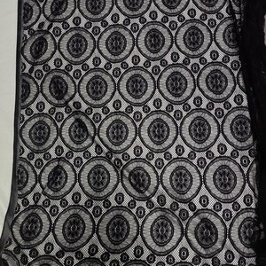 Peut inclure: Un tissu en dentelle noire pr&eacute;sentant un motif g&eacute;om&eacute;trique r&eacute;p&eacute;titif compos&eacute; de grands cercles. Chaque cercle contient un dessin floral central entour&eacute; d'une fine maille. La dentelle noire est superpos&eacute;e sur un fond blanc uni.