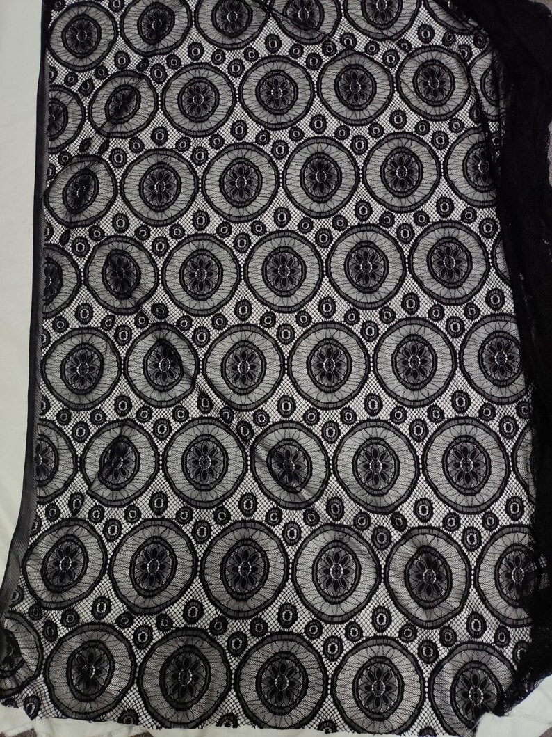Peut inclure: Un tissu en dentelle noire pr&eacute;sentant un motif g&eacute;om&eacute;trique r&eacute;p&eacute;titif compos&eacute; de grands cercles. Chaque cercle contient un dessin floral central entour&eacute; d'une fine maille. La dentelle noire est superpos&eacute;e sur un fond blanc uni.