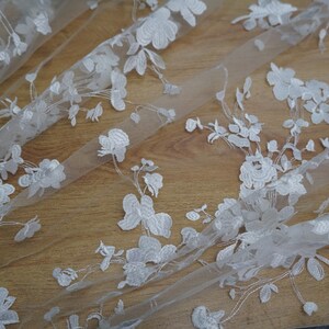 May include: White floral lace fabric featuring a delicate pattern of flowers and leaves. The lace is sheer and has a soft, flowing texture.