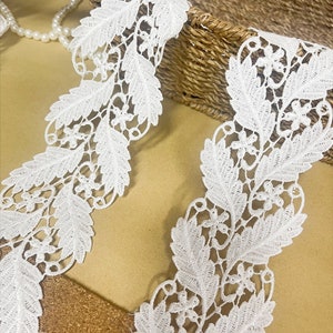 May include: Close-up of white lace trim with a floral and leaf design. The delicate lace is intricately detailed, featuring a repeating pattern of leaves and small flowers. The trim is ideal for sewing and craft projects.