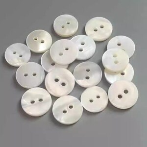 May include: A collection of round, white mother-of-pearl buttons with two holes each. The buttons have a pearlescent sheen and are scattered on a gray surface. These buttons are ideal for sewing and craft projects.